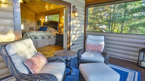 Interior acogedor de una cabaña con una cama, dos sillones, una mesita y vistas al bosque a través de una ventana.