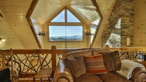 Une chambre confortable aménagée en mezzanine, avec une grande fenêtre, une cheminée en pierre et un plafond en bois.