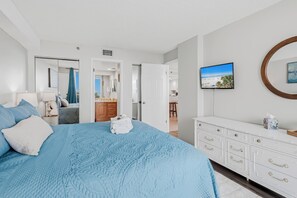 A bedroom with a large bed, a white dresser, a television on the wall, and a door leading to another room.