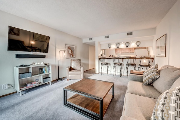 A modern living room with a sofa, coffee table, and a TV. There is a kitchen area with a bar and chairs.