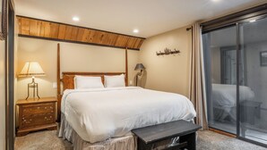 Une chambre à coucher avec un grand lit, une tête de lit en bois, des tables de chevet, un banc et une porte-fenêtre coulissante donnant sur un balcon.