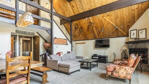 Un vaste espace de vie aménagé dans un style loft, avec un plafond haut, des poutres apparentes, une cheminée, un téléviseur et un coin salon confortable comprenant un canapé et des fauteuils.
