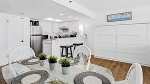 Una cocina moderna con armarios blancos, un refrigerador de acero inoxidable y un comedor con mesa de vidrio y sillas de mimbre.