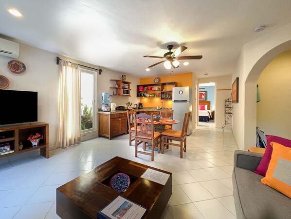 A modern living room with a flat-screen TV, a coffee table with a bowl of blue beads, and a gray sofa with orange and pink cushions. - Quiet apartment with large, private patio, king bed & well-equipped kitchen. (Puerto Morelos)