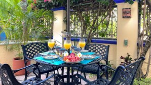 Une table dressée à l’extérieur, sous un parasol bleu, entourée de plantes tropicales et agrémentée d’un mur décoratif.