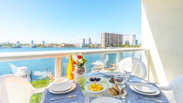 A dining table set with plates, cutlery, and a bottle of orange juice, overlooking a body of water with buildings in the background. - Bright condo with amazing bay views. WIFI + Parking (Sunny Isles Beach)
