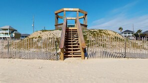 Une structure en bois installée sur la plage, avec un escalier menant à une zone abritée, le tout entouré d’une clôture en bois et avec des maisons en arrière-plan.