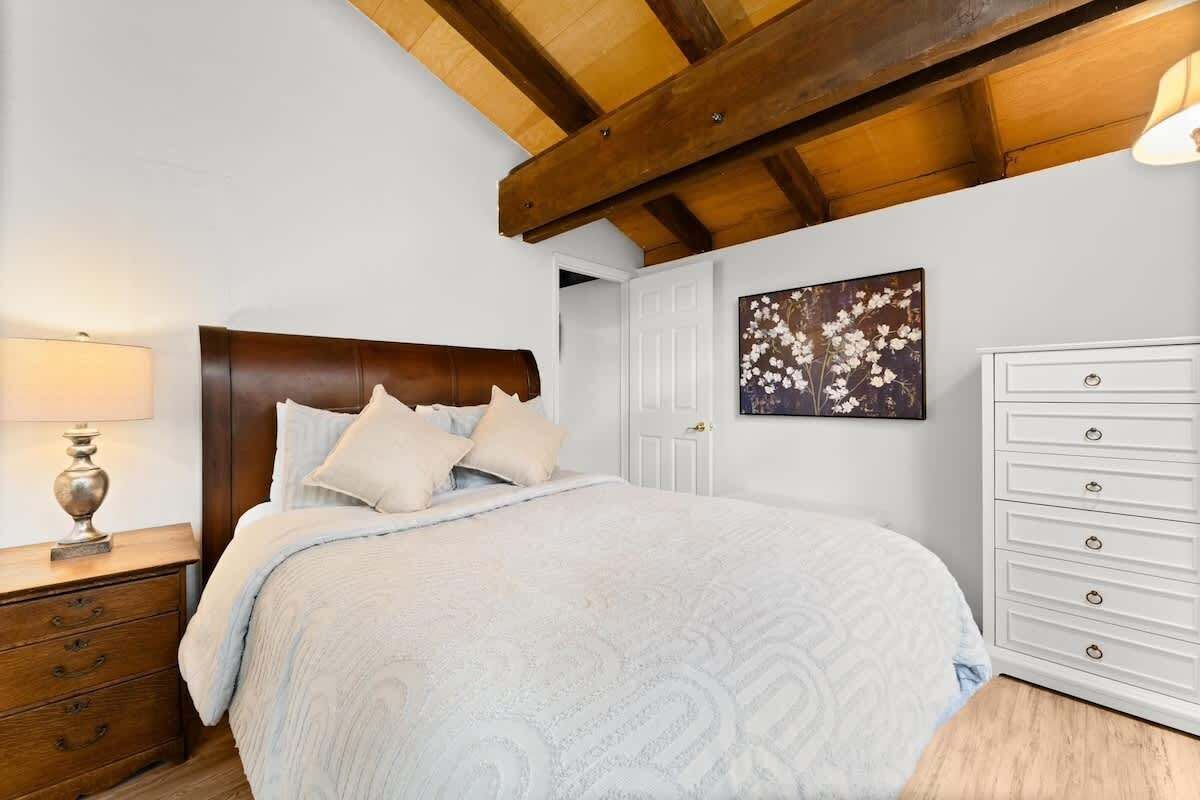 A bedroom with a wooden headboard, a bed with a patterned bedspread, a nightstand with a lamp, a dresser, and a wall-mounted artwork.