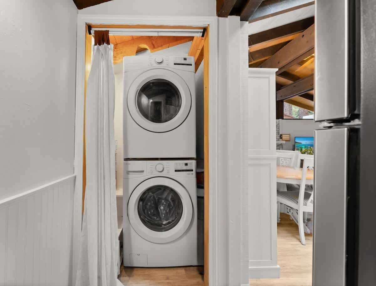 Une buanderie équipée de deux machines à laver superposées, avec un plafond en bois et un coin repas visible à travers une porte ouverte.