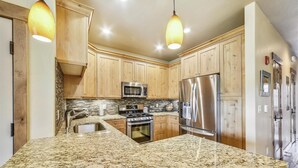 A kitchen with granite countertops, wooden cabinets, stainless steel appliances, and pendant lighting.