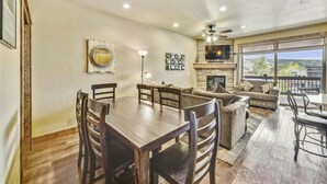 A spacious living room with a fireplace, a dining area with a wooden table and chairs, and a large window offering a view of the outdoors.