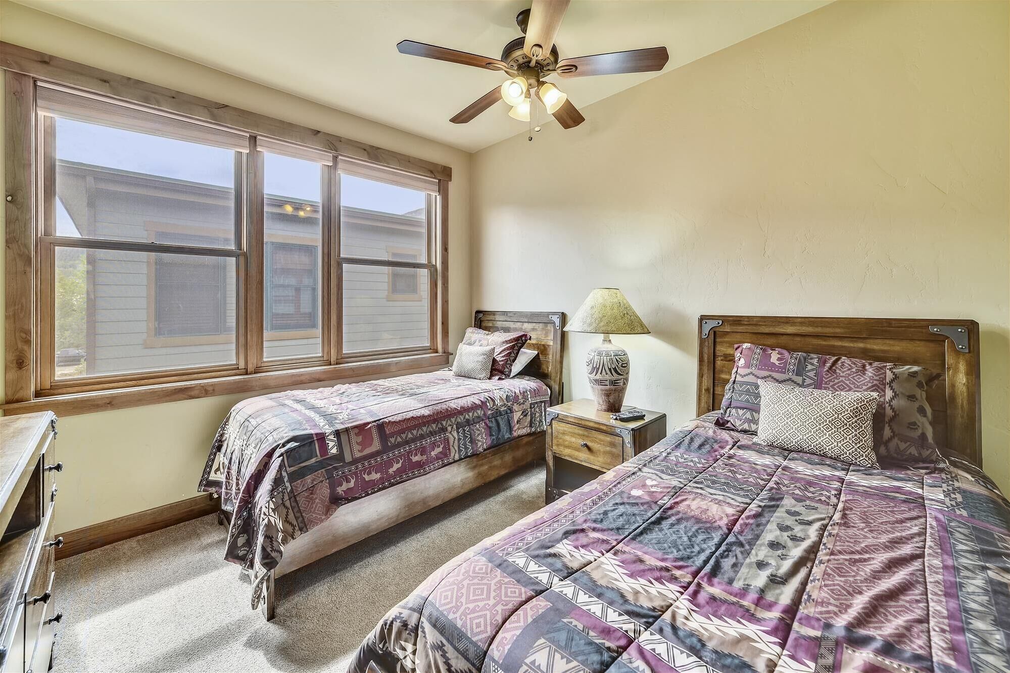 A bedroom with two beds, a ceiling fan, a nightstand with a lamp, and a window with blinds.