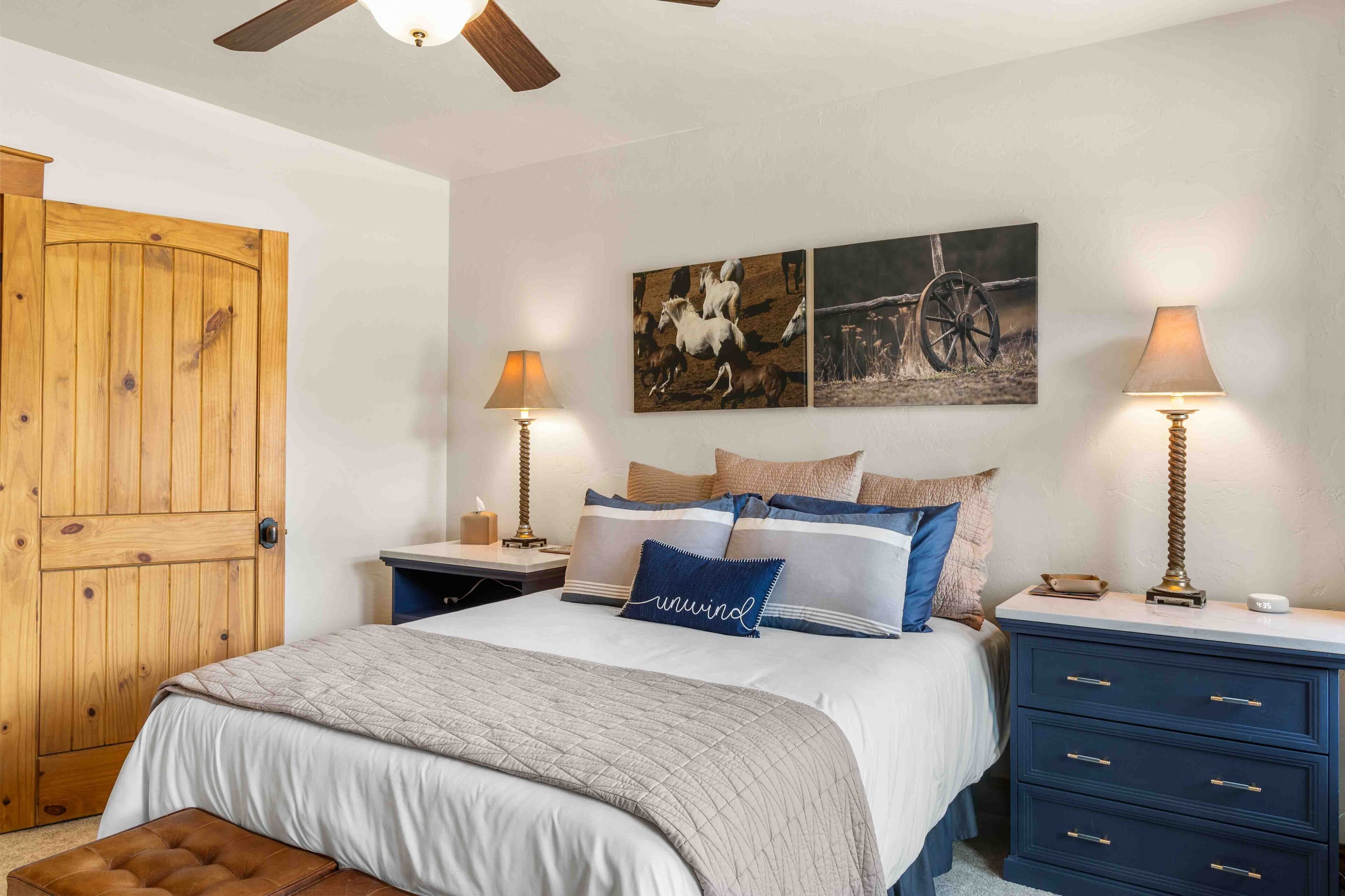 A bedroom with a wooden door, a bed with a quilt, two bedside tables with lamps, and wall art.