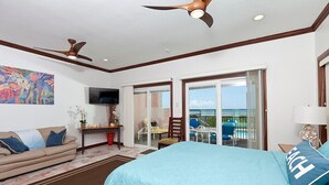 Habitación de hotel con cama, sofá, televisión, balcón y vistas a la piscina, además de ventiladores de techo.