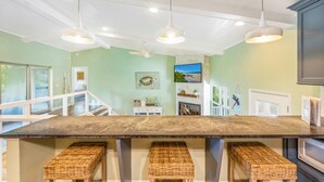 Una cocina amplia con una isla grande, taburetes de mimbre, iluminación colgante y vistas al exterior.