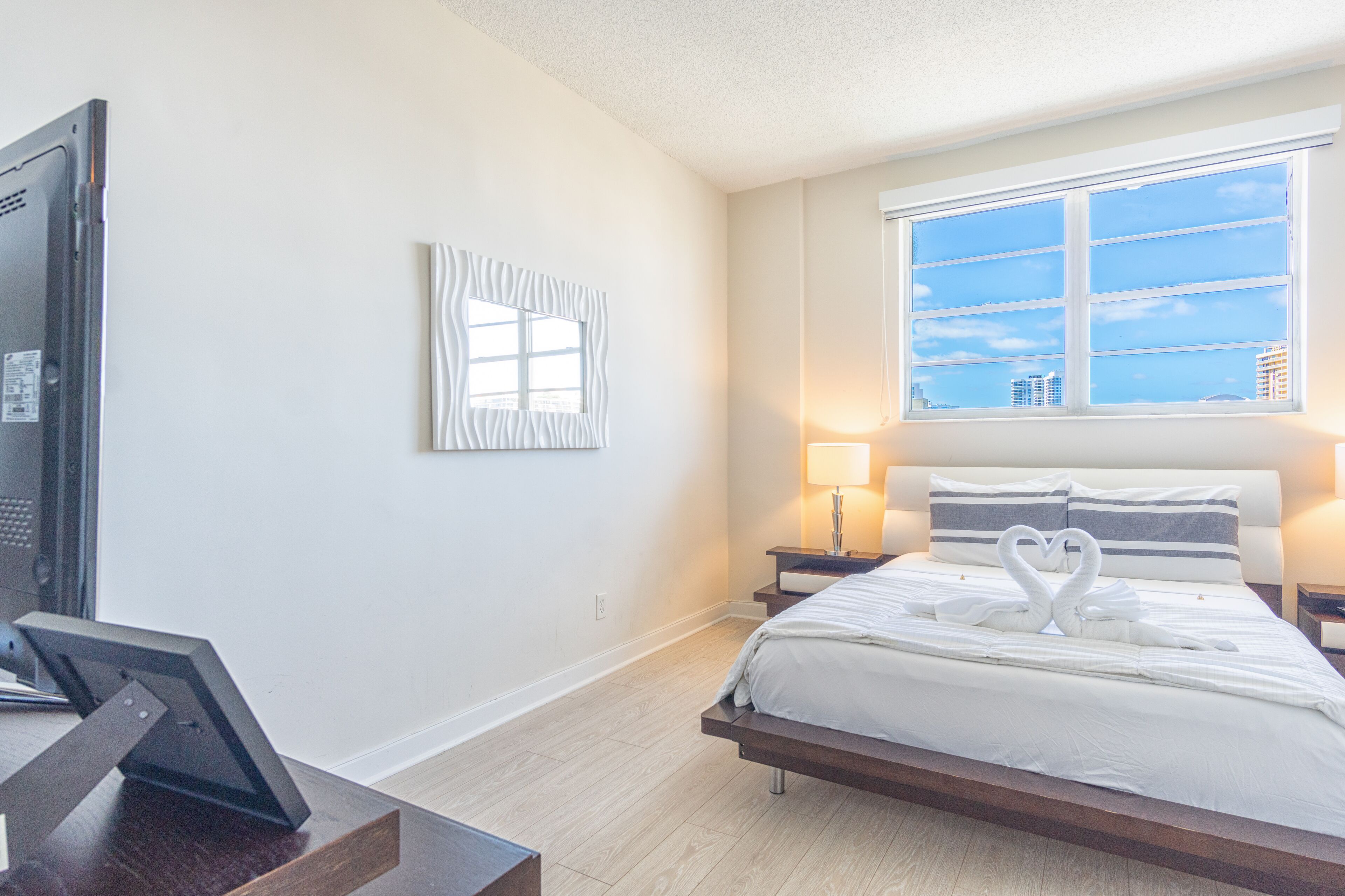 A modern hotel room with a large bed, a desk with a laptop, a window with a city view, and a wall-mounted artwork.