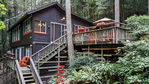 Una cabaña de madera de dos pisos con un balcón y escaleras, rodeada de un bosque frondoso.