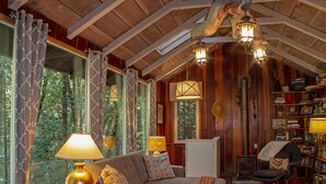 Un interior de cabaña acogedor con techo de madera, ventanas amplias, un sofá, una lámpara y una estantería.