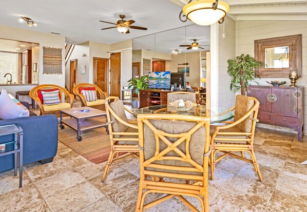 A spacious living area with a glass-top dining table surrounded by wicker chairs, a blue sofa, and a television in the background.