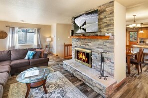 A living room with a stone fireplace, a television mounted above it, and a dining area visible in the background.