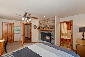 A bedroom with a fireplace, a ceiling fan, a bed, a desk, and a door leading to another room.
