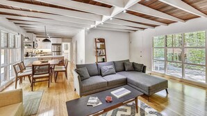 Un espacio habitable amplio con un sofá seccional gris grande, una mesa de comedor de madera y sillas, una mesa baja con libros, y una cocina al fondo.
