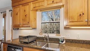 A kitchen with wooden cabinets, granite countertops, stainless steel sinks, and a window with a view of trees. - Cliffside Romantic Mountain Getaway - Hot Tub, Fireplace, Pet Friendly, and View (Ferguson)