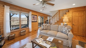 A spacious living room with a wooden floor, a ceiling fan, a large window with a view of the outdoors, a sofa with cushions, and a staircase leading to another level. - Cliffside Romantic Mountain Getaway - Hot Tub, Fireplace, Pet Friendly, and View (Ferguson)