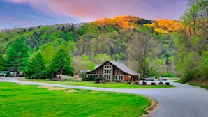 A rustic cabin with a large porch, surrounded by lush green fields and a forested hill in the background. - Cliffside Romantic Mountain Getaway - Hot Tub, Fireplace, Pet Friendly, and View (Ferguson)