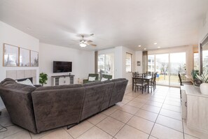 A modern living room with a grey sofa, a flat-screen TV, a ceiling fan, and a dining area with a table and chairs.