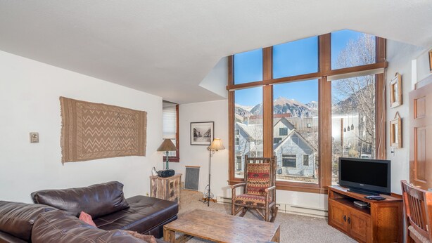 A living room with a leather sofa, a wooden coffee table, a television on a wooden stand, and large windows offering a mountain view. - Classic Ski Condo, Steps to Chair 7 and on shuttle route (Telluride)