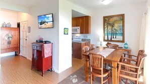 A dining area with a wooden table and chairs, a kitchen in the background, and a painting on the wall. - Bright, Airy Condo w/Epic View! Wood Floors, Lanai, Full Kitchen, WiFi–Kaha Lani 225 (LIHUE)