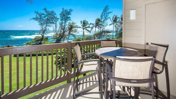 A balcony with a view of the ocean, a round table, and wicker chairs. - Bright, Airy Condo w/Epic View! Wood Floors, Lanai, Full Kitchen, WiFi–Kaha Lani 225 (LIHUE)