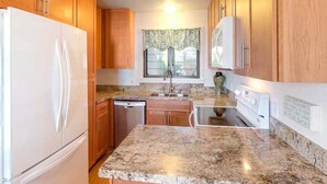 A kitchen with wooden cabinets, a granite countertop, a refrigerator, a dishwasher, and a window with a patterned curtain. - Bright, Airy Condo w/Epic View! Wood Floors, Lanai, Full Kitchen, WiFi–Kaha Lani 225 (LIHUE)