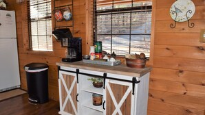 A wooden cabin interior with a kitchen counter, a coffee maker, a trash can, and a window with blinds.