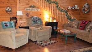 A cozy living room with a wooden staircase, a sign reading "Friends Gather Here," and a variety of seating options including a sofa and armchairs.