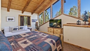 A bedroom with a large bed, a wooden dresser, a television, and a window with a view of trees. - Luxury Log Style Home in High Timber Ranch Estates! 1 Block from National Forest! (Big Bear City)