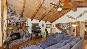 A cozy living room with a stone fireplace, wooden beams, a ceiling fan, and a large gray sectional sofa. - Luxury Log Style Home in High Timber Ranch Estates! 1 Block from National Forest! (Big Bear City)