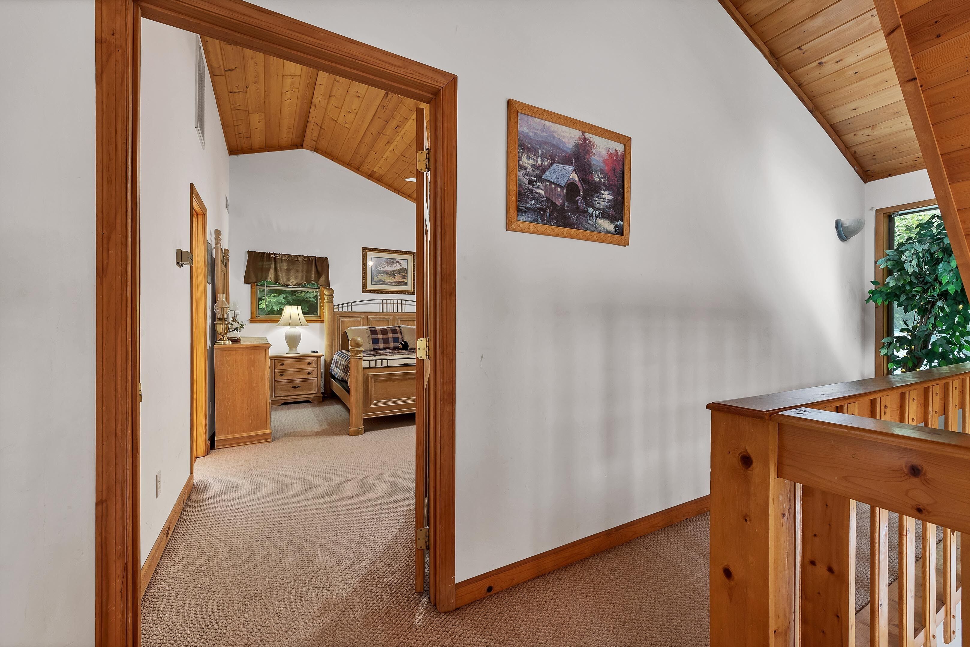 A room with a wooden ceiling, a painting on the wall, a bed with a headboard, and a window with a view of greenery.