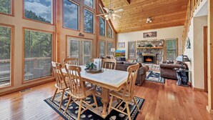 Un vaste salon doté d’un plafond en bois, de grandes fenêtres, d’une cheminée et d’un coin repas avec des chaises et une table en bois.