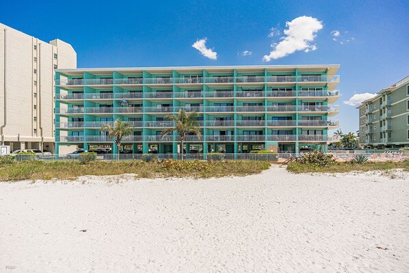 Et flereetagers hotel med balkoner, beliggende på en sandstrand med palmer.