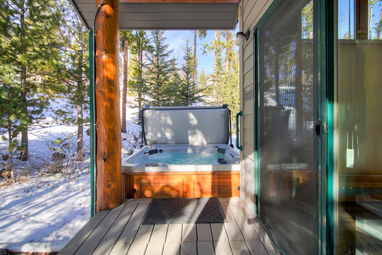 A hot tub on a wooden deck with a glass door, surrounded by trees and snow.