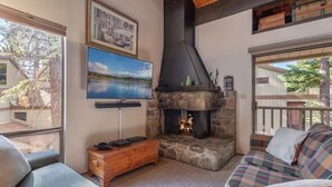 Ein gemütliches Wohnzimmer mit Kamin aus Stein, einem an der Wand montierten Fernseher und Blick ins Freie.