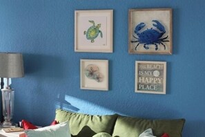 A room with a blue wall, a framed turtle and crab artwork, a framed seashore quote, and a green couch with red cushions.