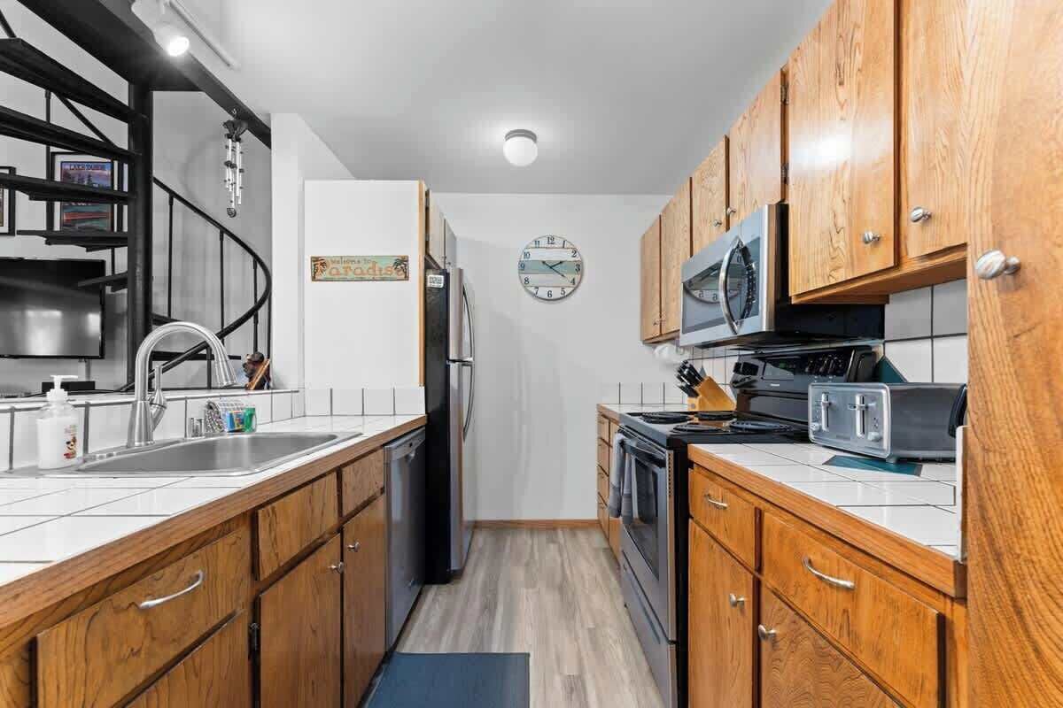 A compact kitchen with wooden cabinets, a stainless steel sink, a microwave, and a clock on the wall.