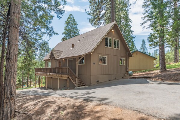 A two-story cabin with a covered porch and a garage, surrounded by tall pine trees.