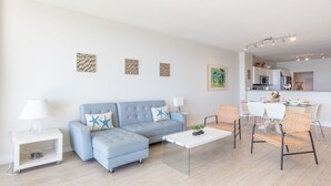 Un salon moderne avec un canapé, une table basse et des fauteuils. On aperçoit un coin cuisine au fond.