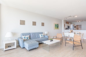 Un salon moderne avec un canapé, une table basse et des fauteuils. On aperçoit un coin cuisine au fond.