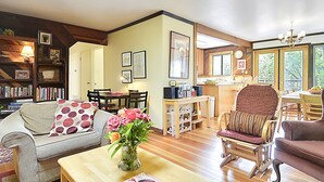 A cozy living room with a sofa, armchair, and a coffee table with a vase of flowers. There is a bookshelf, a dining table with chairs, and a kitchen area visible in the background. - Boathouse ~ On River, Deck, Steps to Beach, Family  Dog Friendly! (Guerneville)