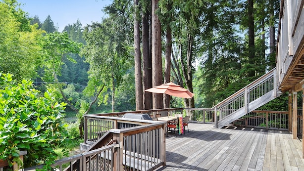A wooden deck with a grill and outdoor furniture, surrounded by tall trees. - Boathouse ~ On River, Deck, Steps to Beach, Family  Dog Friendly! (Guerneville)